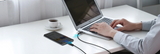 A person using a silver laptop on a white desk, with a smartphone charging via a black cable placed beside it. A cup of coffee sits nearby in a bright indoor setting.