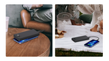 A black portable power bank with a blue LED light glow, shown in two lifestyle settings: on a wooden table next to a person, and outdoors on a picnic blanket charging a smartphone.