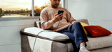 Man sitting on a couch by a window, using a smartphone with a scenic view outside.