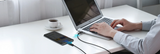 A person typing on a silver laptop at a white desk, with a smartphone connected via a charging cable on the table next to a cup of coffee.